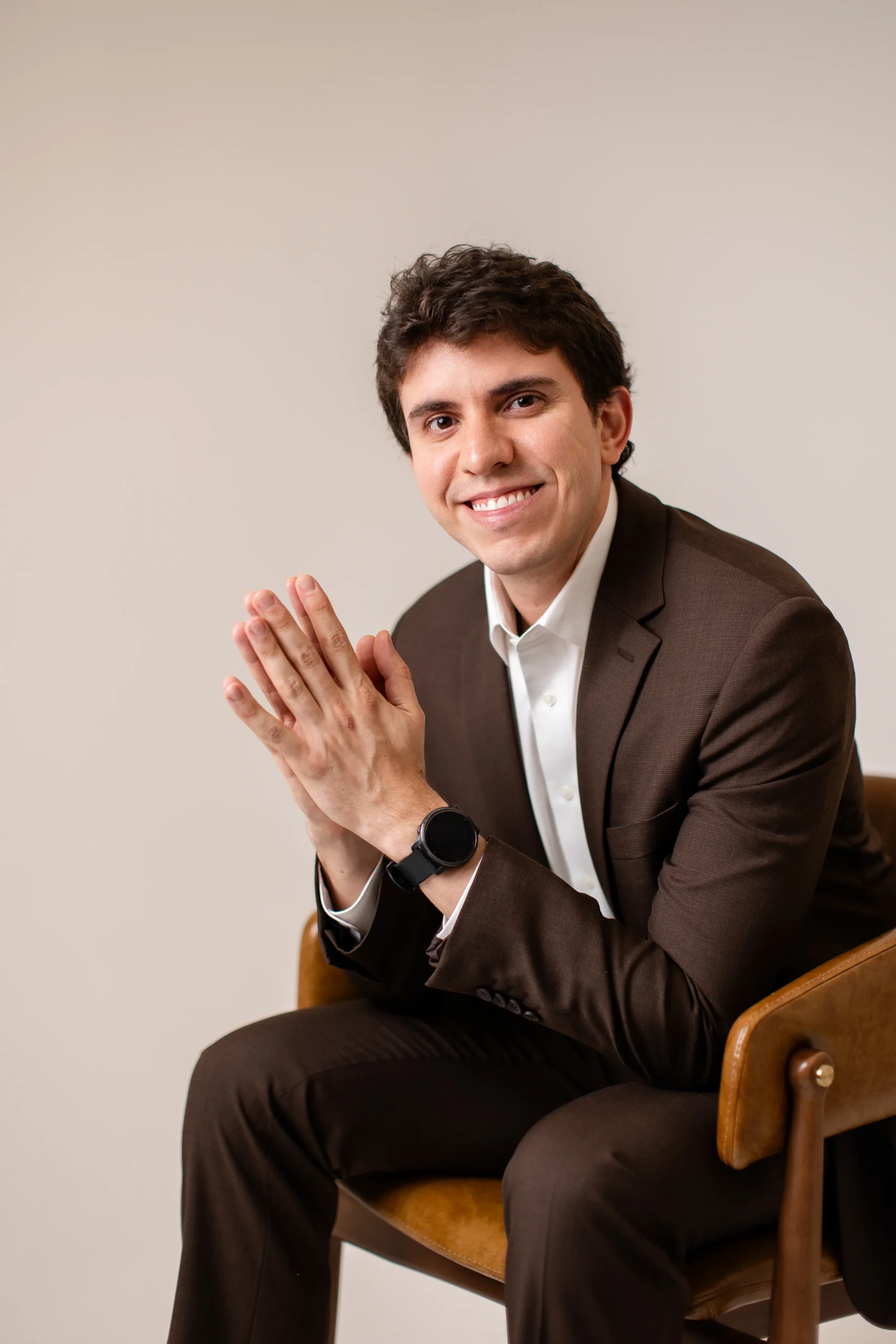 Dr. Matheus Massahud sorrindo, sentado de terno marrom, com as mãos unidas