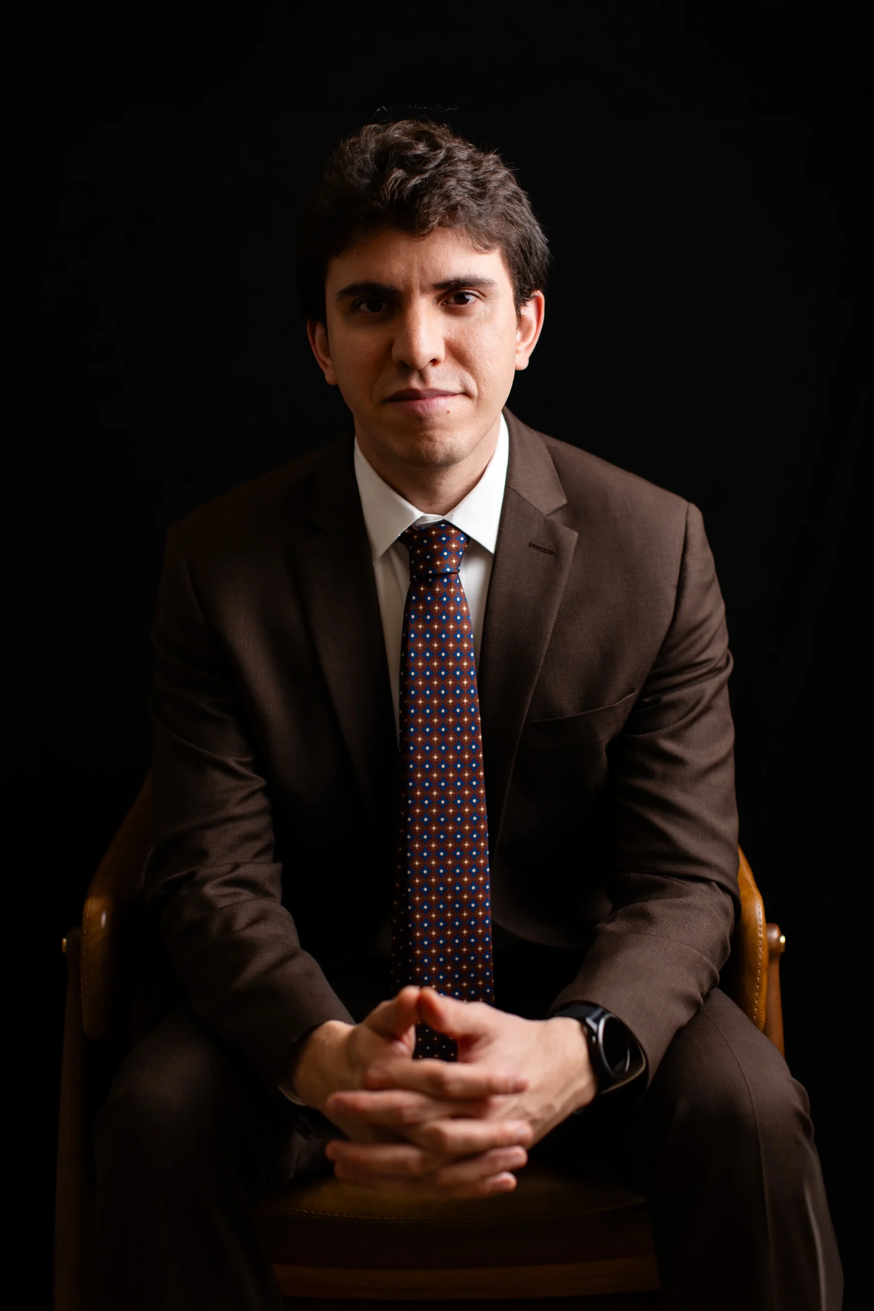 Dr. Matheus Massahud sentado, de terno, com as mãos unidas, em retrato com fundo escuro.
