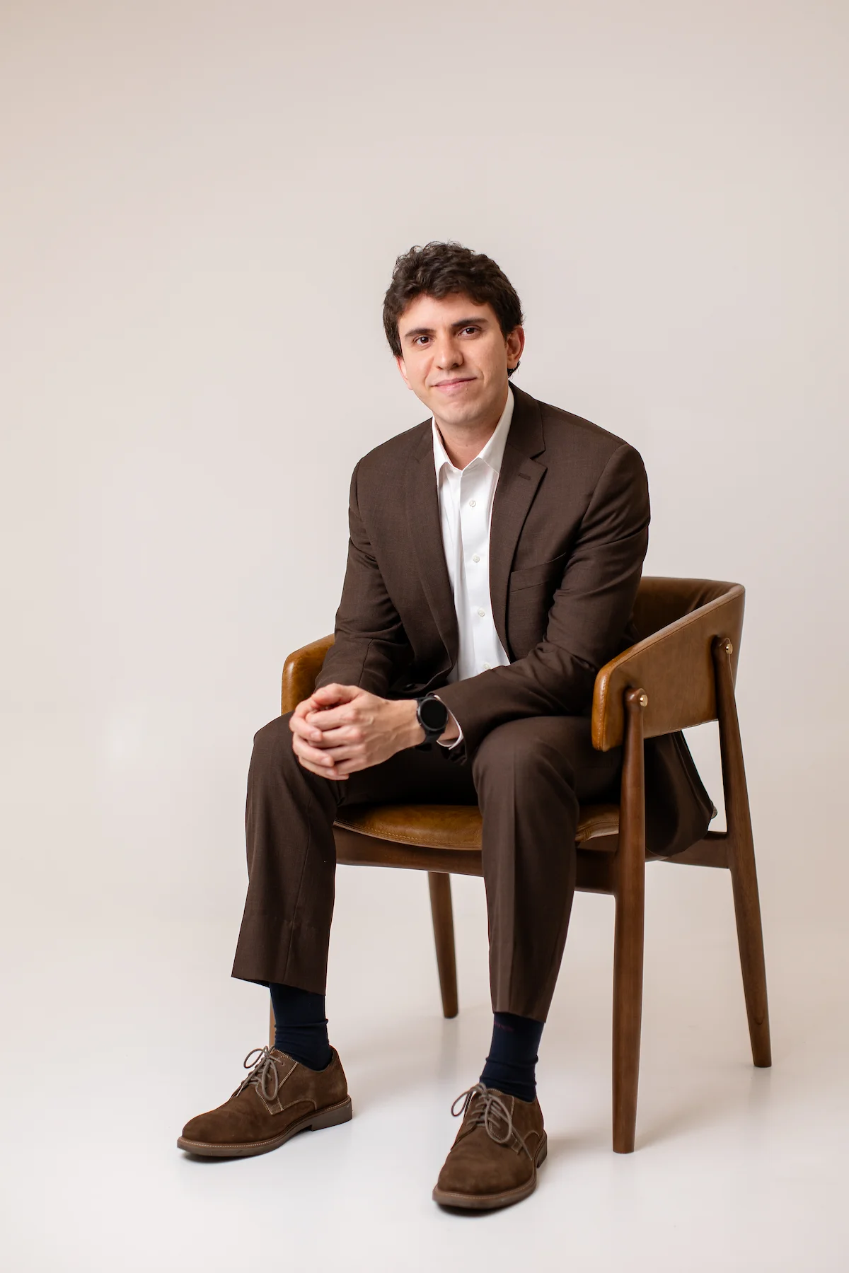 Dr. Matheus Massahud sentado em uma cadeira, de terno marrom, em retrato profissional com fundo claro