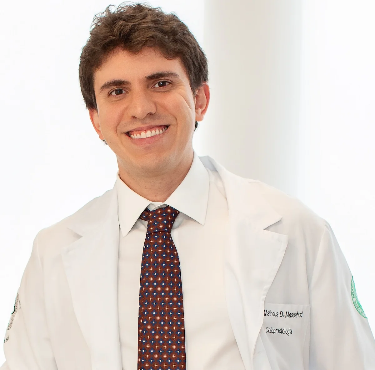 Dr. Matheus Massahud sorrindo, usando jaleco branco, em retrato profissional.