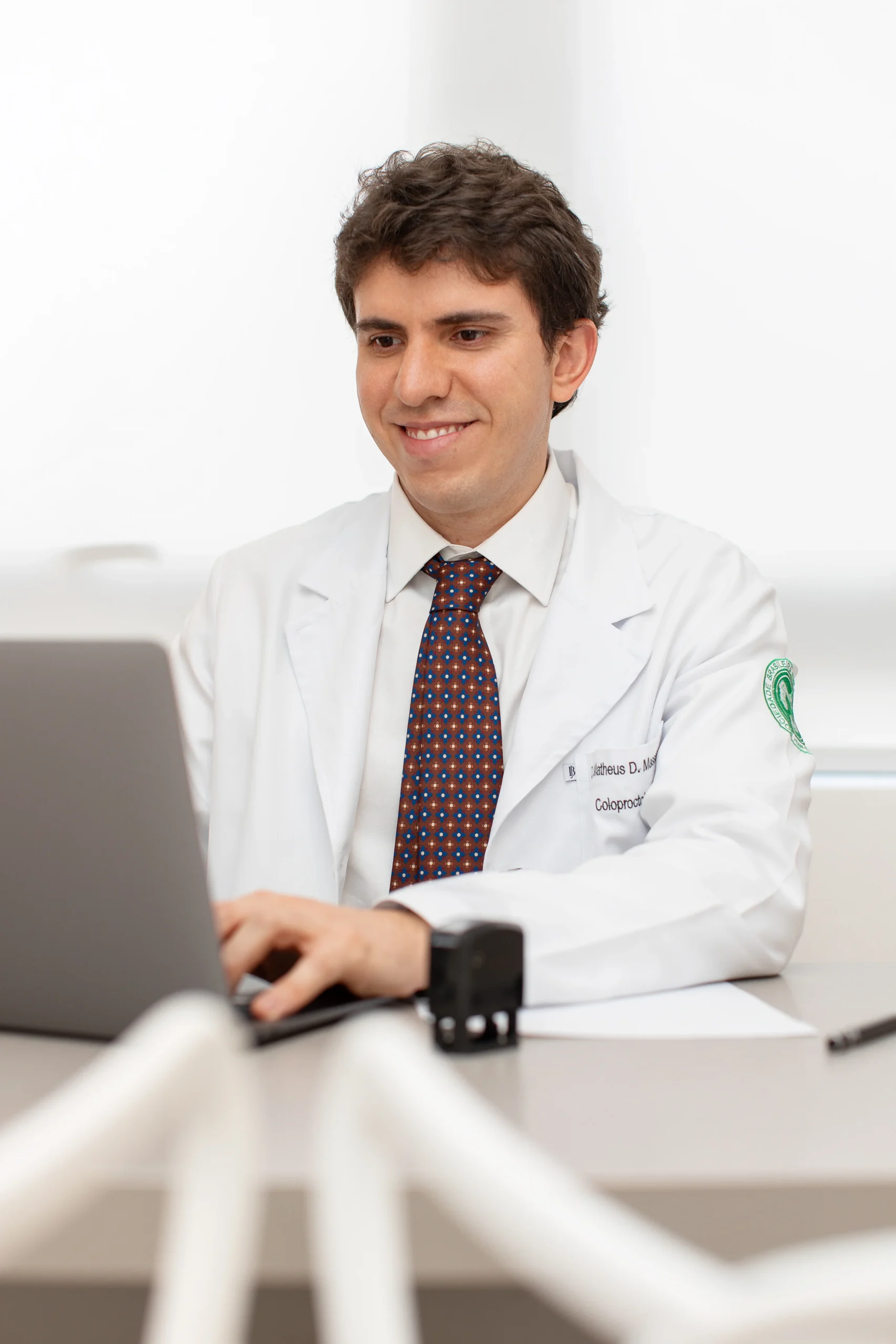 Dr. Matheus Massahud sorrindo, de jaleco, sentado em seu consultório