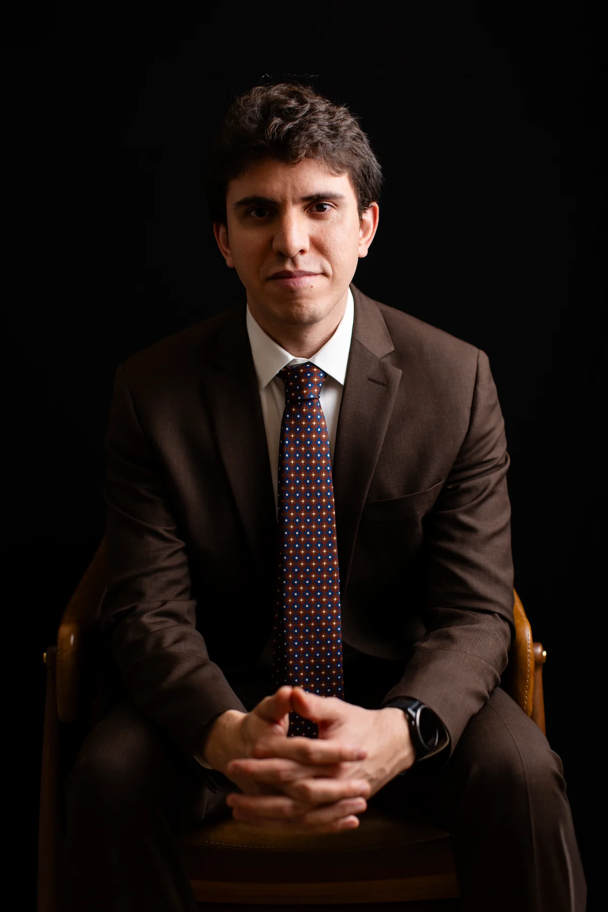 Dr. Matheus Massahud sentado, de terno, com as mãos unidas, em retrato com fundo escuro.