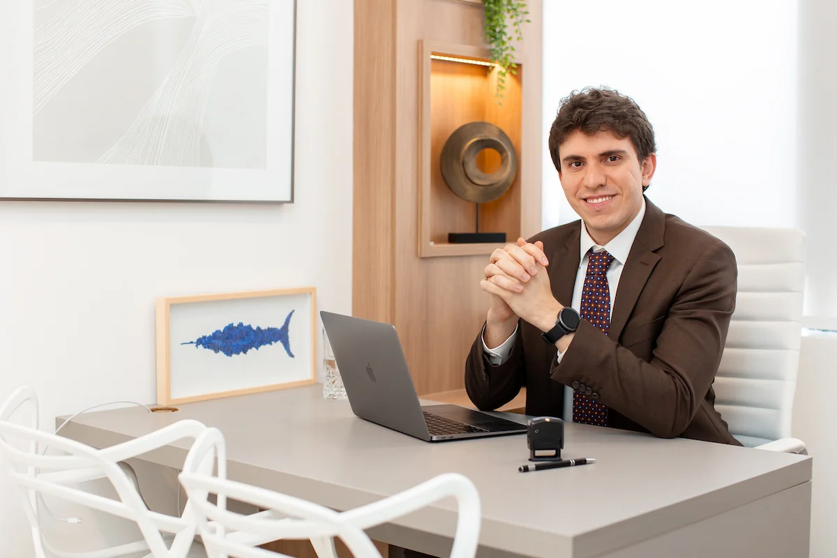 Dr. Matheus Massahud sentado à mesa do consultório, com as mãos unidas e um computador à sua frente.