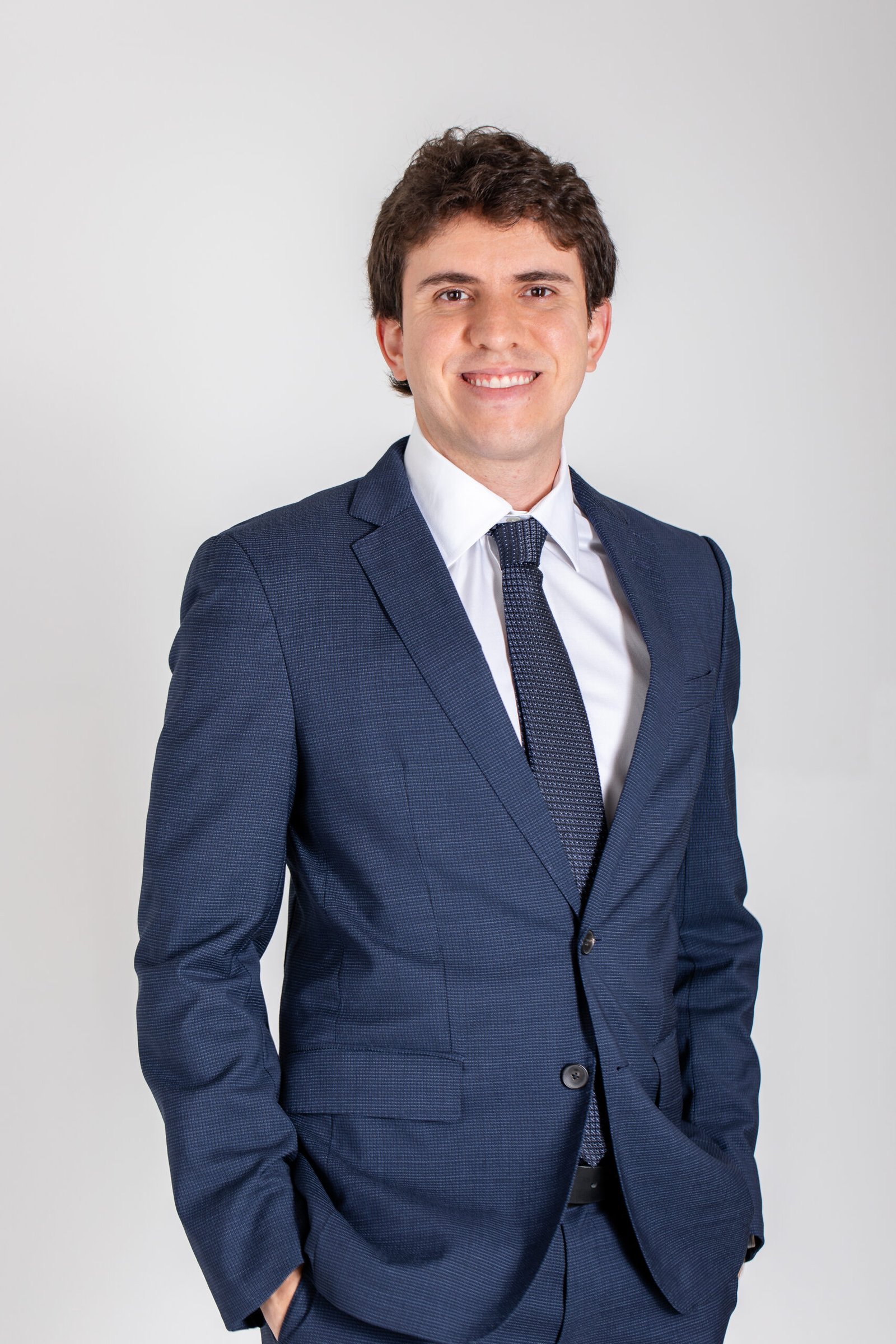 Dr. Matheus Massahud sorrindo, de terno azul, em retrato profissional com fundo claro.