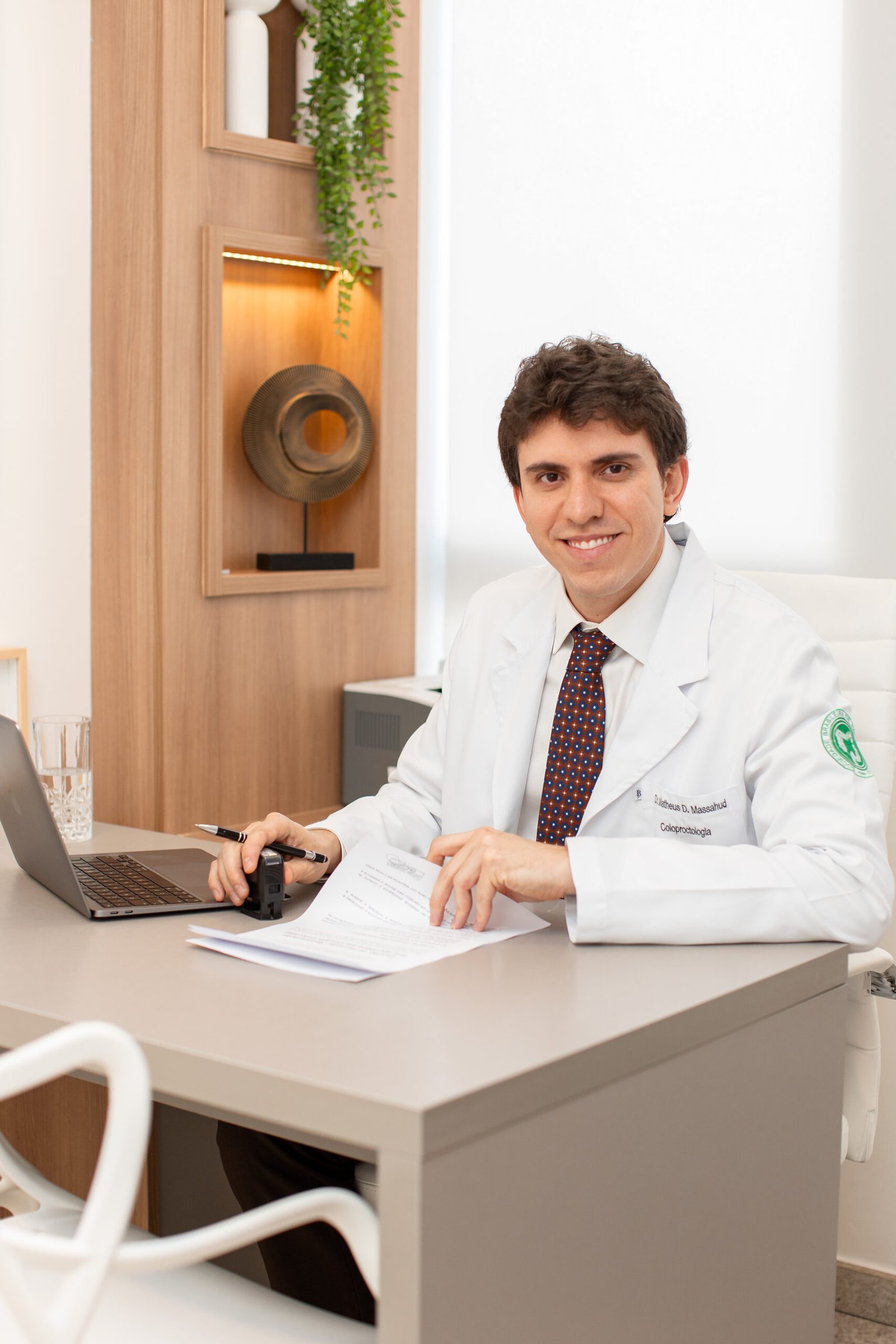 Dr. Matheus Massahud de jaleco, sentado à mesa do consultório, preenchendo um documento.