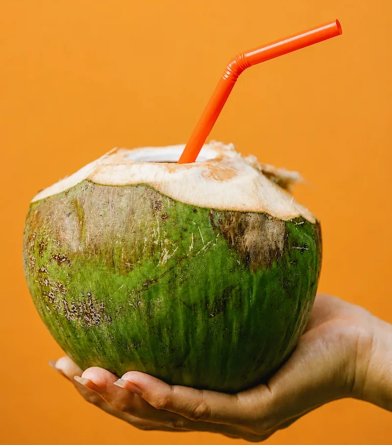 Mão segurando um coco verde aberto, com canudo laranja, diante de fundo laranja liso