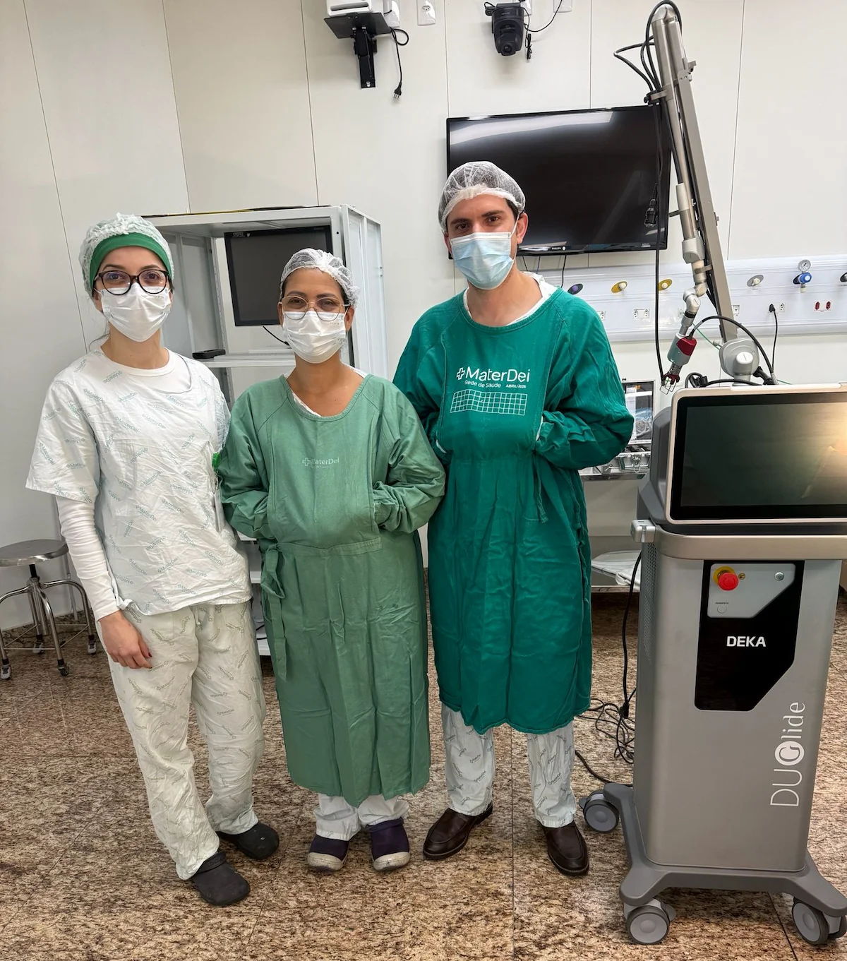 Equipe médica em centro cirúrgico, usando roupas hospitalares, toucas e máscaras, ao lado de equipamento cirúrgico a laser.